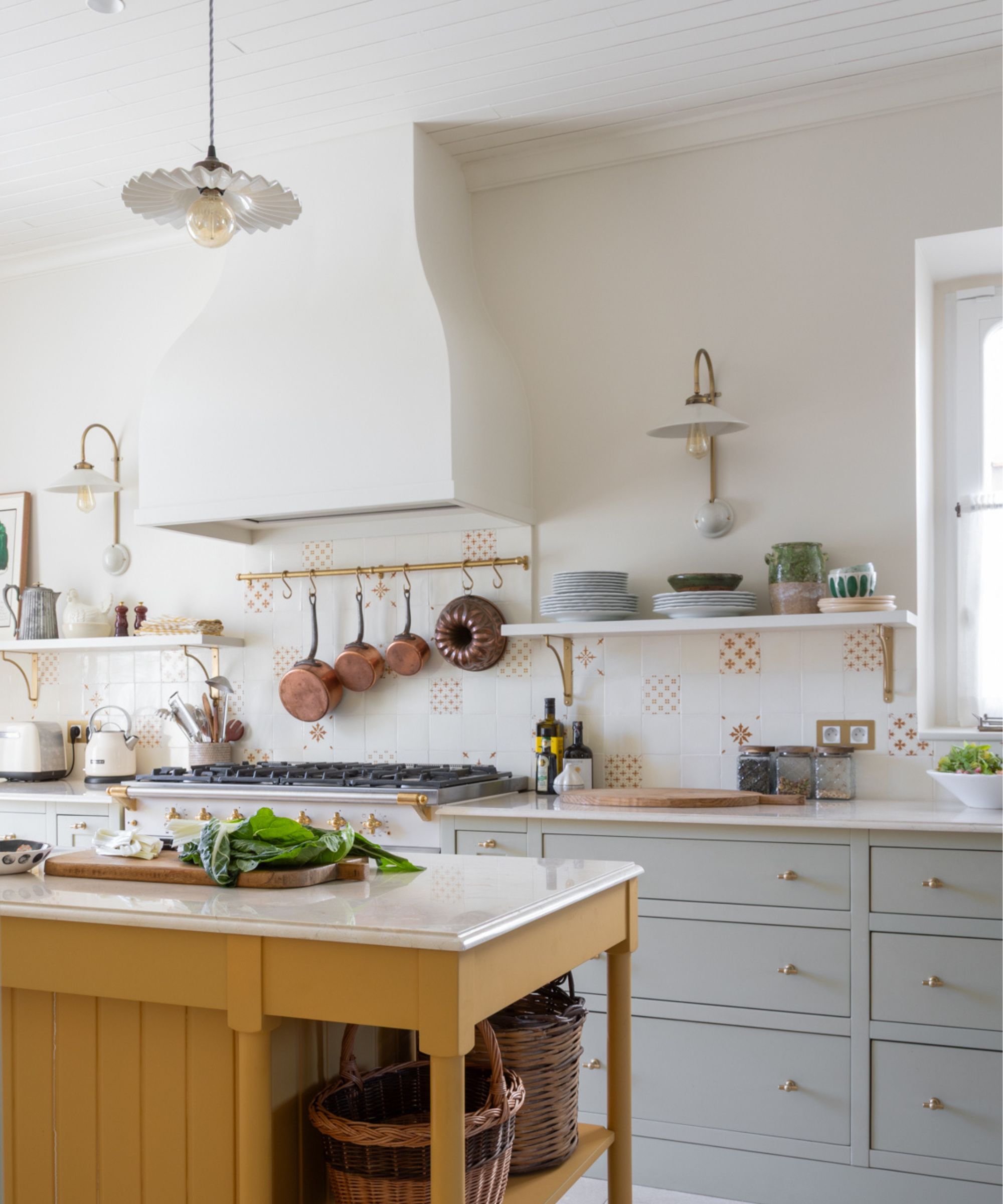 A bright kitchen with pale gray cabinetry, open shelving, a white range hood, brass fixtures, and a central island in mustard yellow