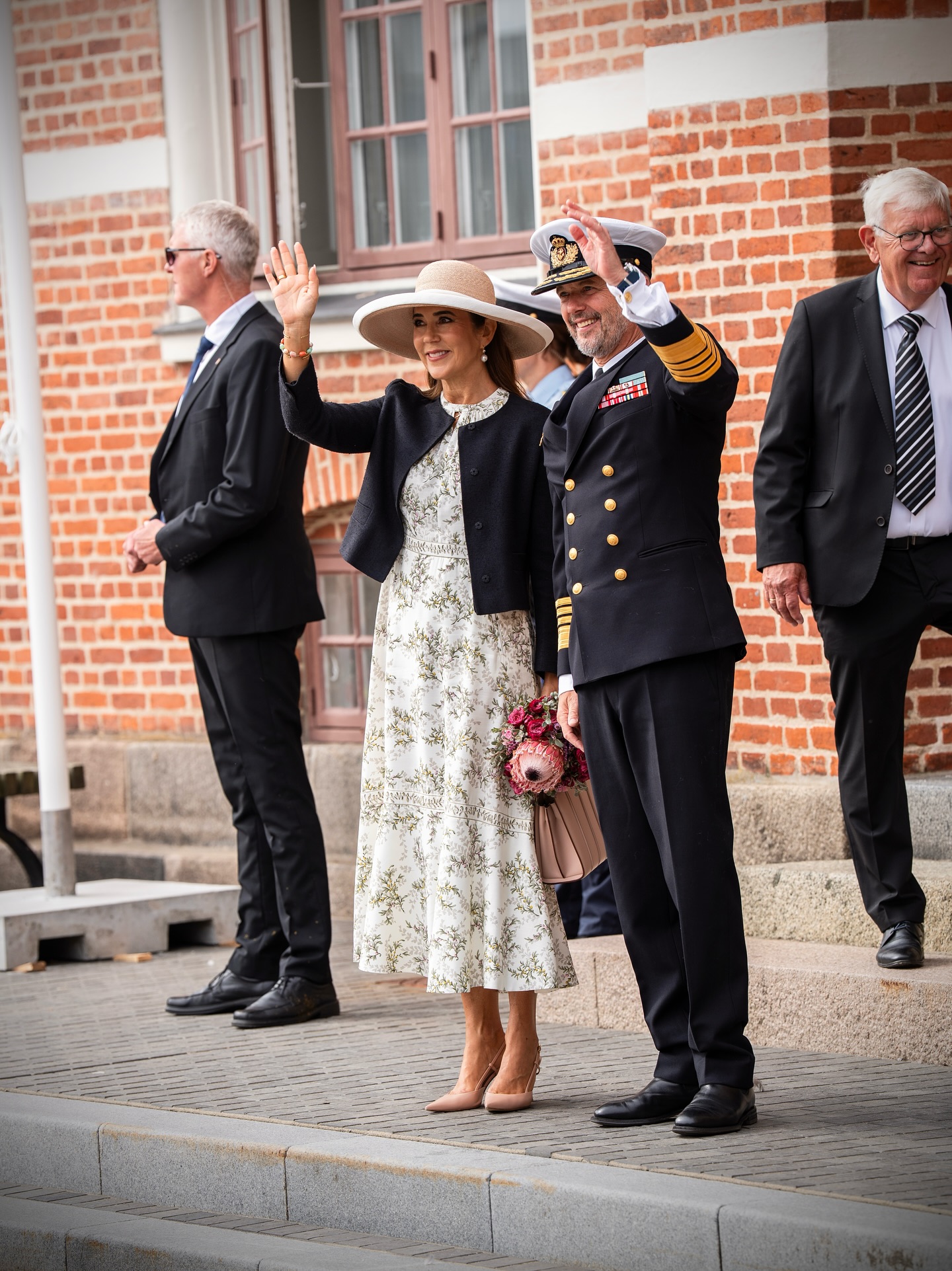 Queen Mary wearing a white dress and black jacket waving next to King Frederik wearing a naval uniform