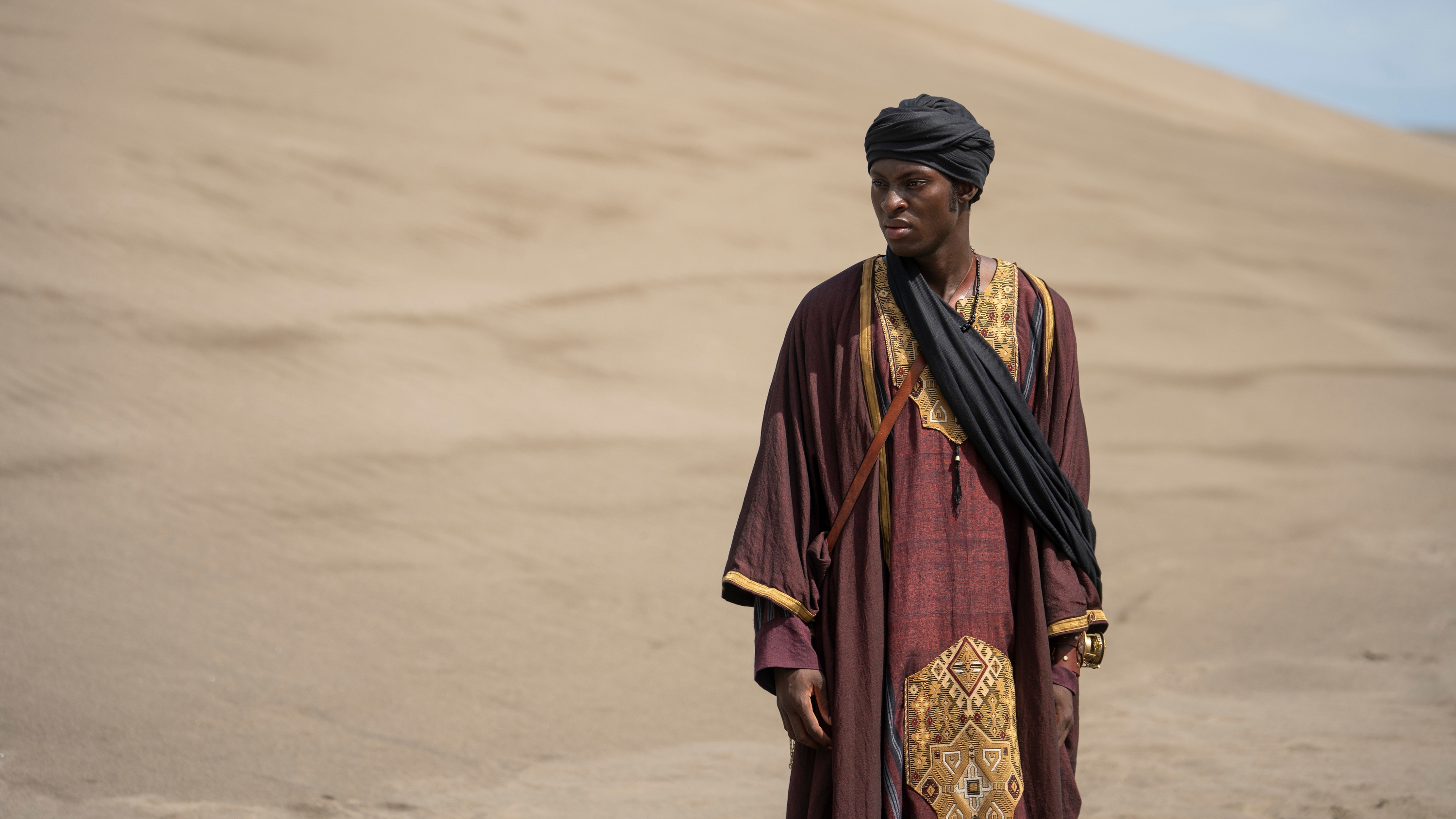 Ernest Kingsley Junior as Wash Black in a desert wearing a maroon robe with a gold design and a black turban.