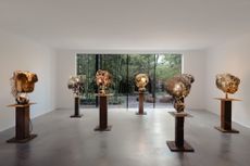 A series of sculptural lights stands on wooden podiums in a gallery with a floor-to-ceiling window giving to a garden.