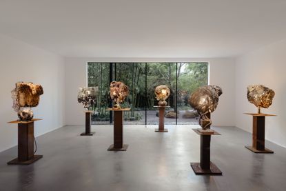 A series of sculptural lights stands on wooden podiums in a gallery with a floor-to-ceiling window giving to a garden.