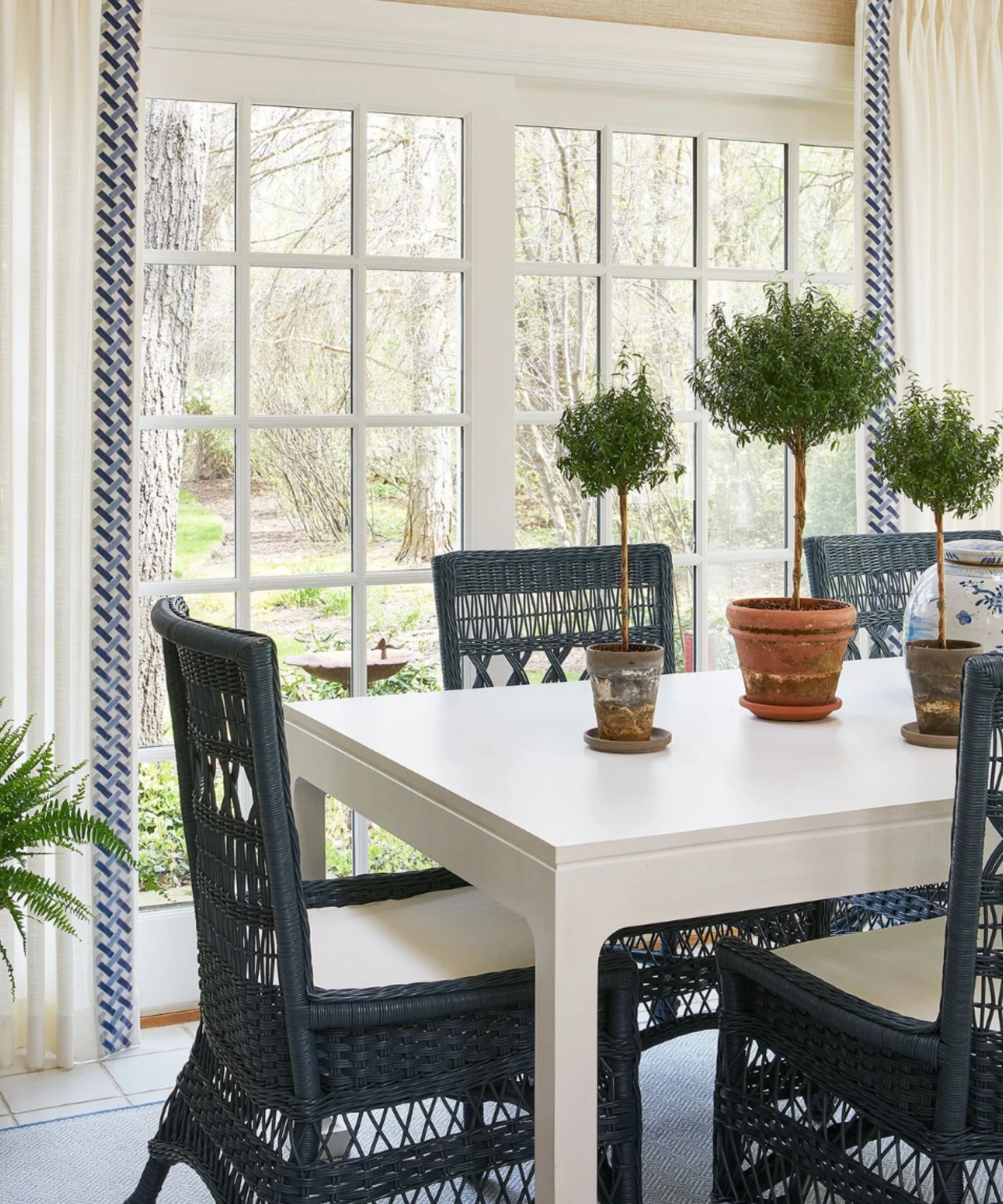 A bright sunroom with off-white curtains finished with blue geometric trim, a white dining table, navy woven dining chairs and large windows overlooking the garden.