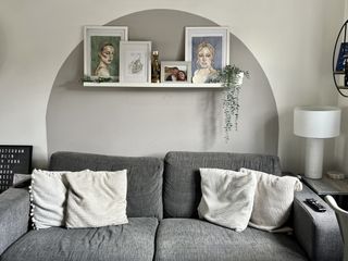 A dark grey sofa with cream cushions, in front of a wall with a grey painted arch, and a decorated shelf with paintings and plants on.