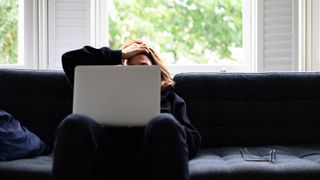 Person slumps over a laptop on their couch looking stressed