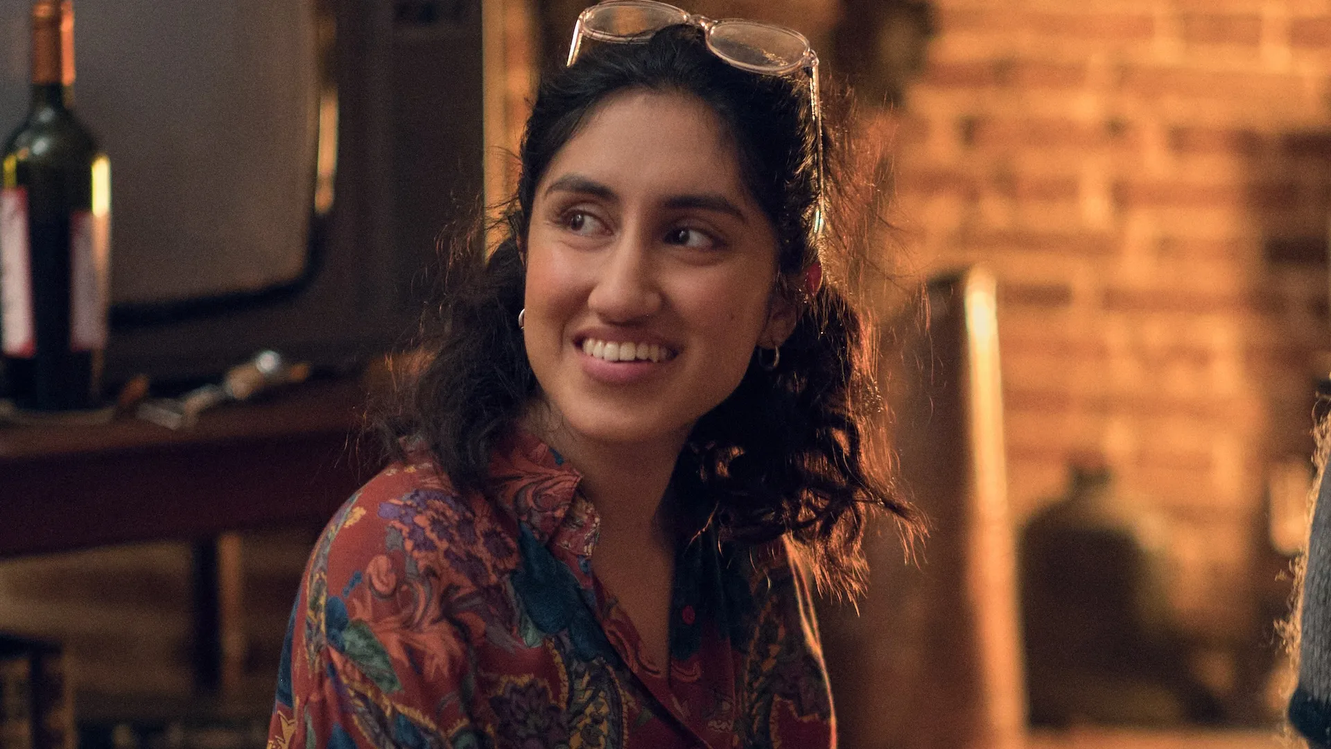 Emma played by Ambika Mod smiles up at an unknown figure with her glasses on her head. A bottle of wine and brick wall are behind her.