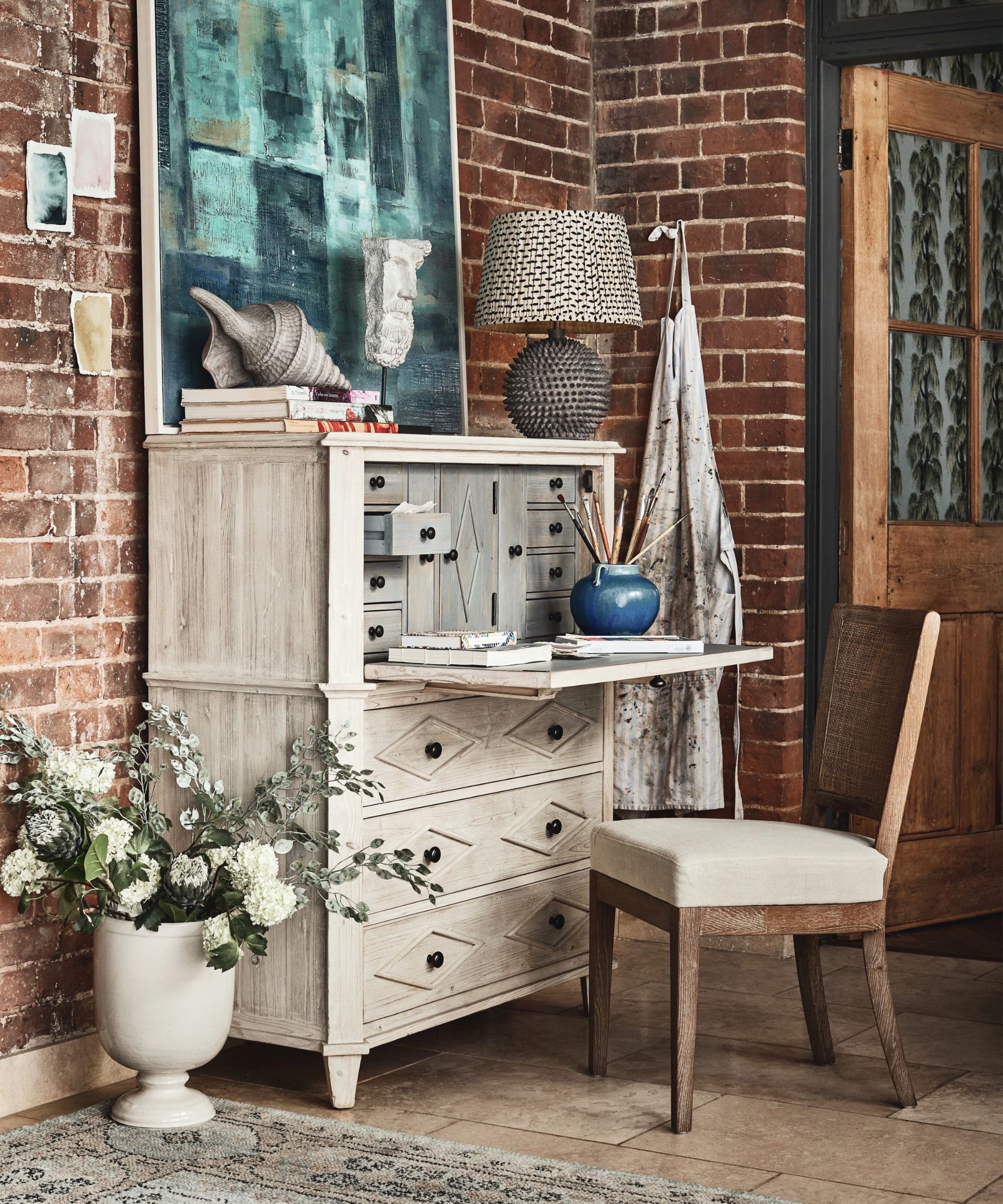 Rustic home workspace with a distressed wood bureau desk, exposed brick wall, sculptural decor, patterned table lamp, and a cosy cushioned chair on tiled flooring.