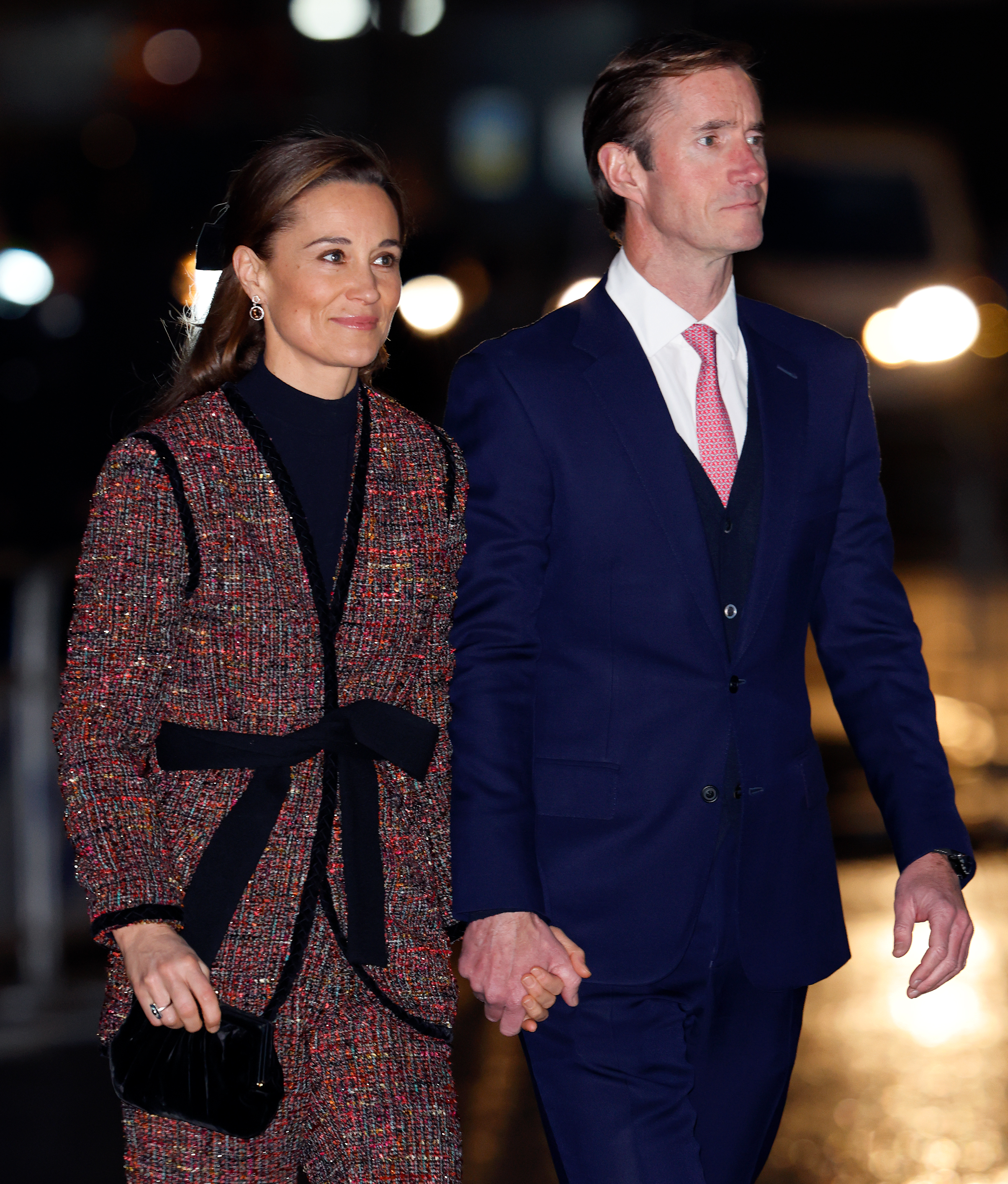 Pippa Middleton wearing a tweed suit with a black bow, holding hands with husband James Matthews in a blue suit