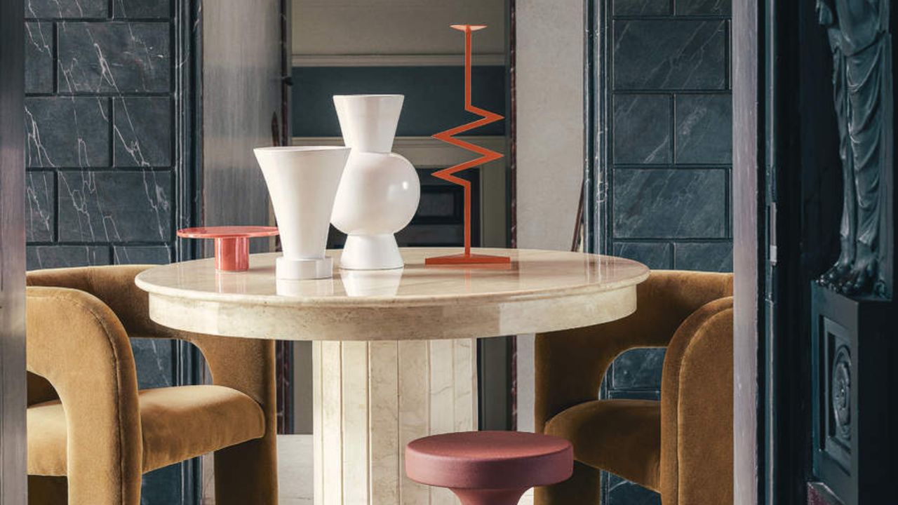 Image of a dining room that has slate walls. There is a round white marble dining table in the center with two ochre yellow, velvet chairs around it and a pink stool. There are two white vases and an abstract, orange candle stick holder.