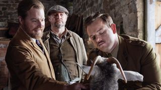 Samuel West in a brown vet's coat as Siegfried and Callum Woodhouse in an army uniform as Tristan treat a goat while Nicholas Asbury in a cap and jacket as Farmer Biggins looks on in All Creatures Great and Small.