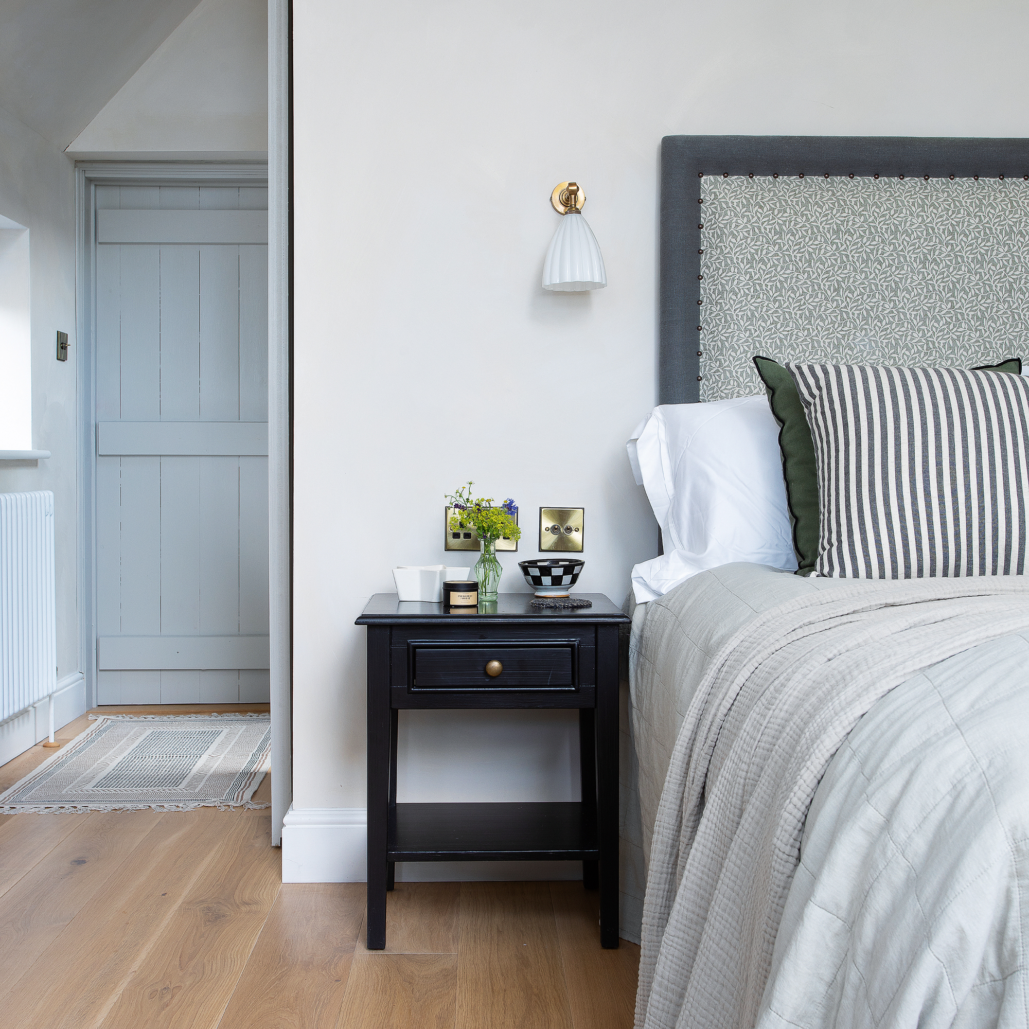 A bedroom with an upholstered headboard, wooden flooring, and a striped black and white cushion on the bed