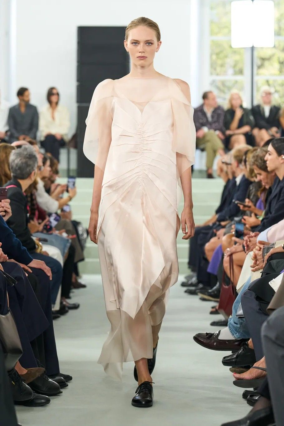 A model dressed in a layered dress in cream with a silk body and see-through, tulle shoulders walks the runway, surrounded by people dressed in formal clothing in the audience.