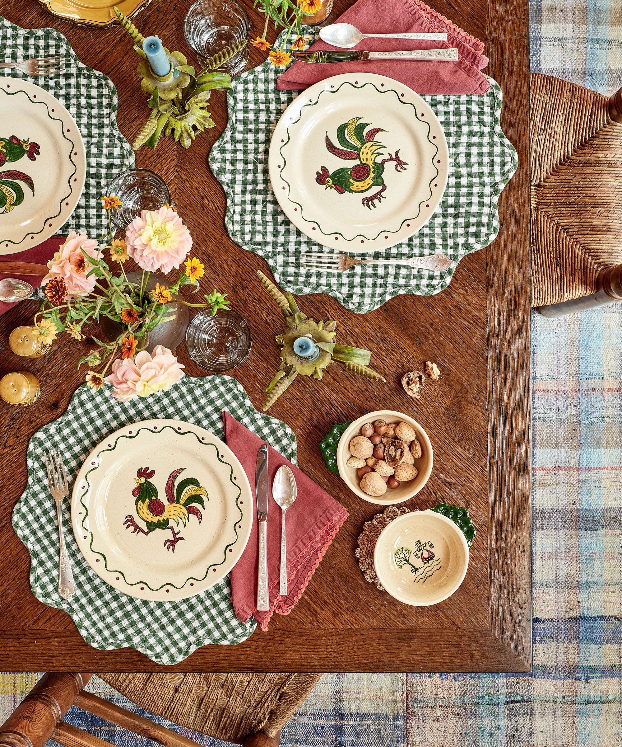 Rustic wooden table with gingham placemats, pink napkins, and painted plates with roosters on, a vase of flowers in the centre, and wicker dining chairs tucked under the table, with a plaid carpet.