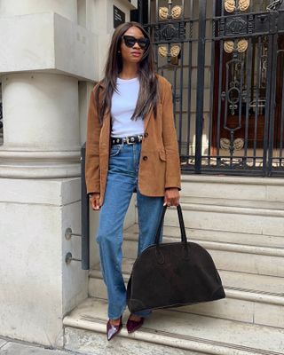 British influencer Lorna Humphrey poses in oversize black sunglasses, a tan suede blazer, white T-shirt, studded black belt, relaxed straight-leg jeans, The Row suede bag, and patent burgundy pumps