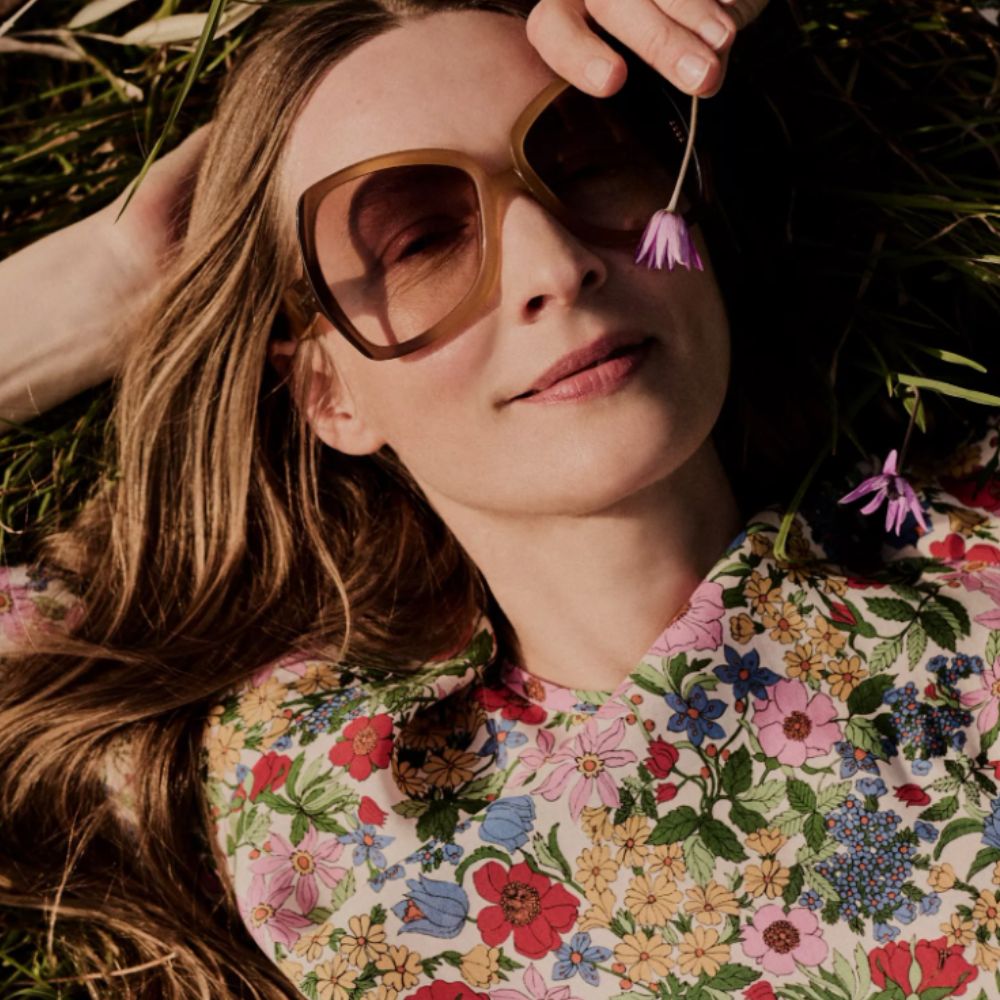 Woman lying on grass and dressed in floral dress, brown sunglasses, smelling pink flower. 