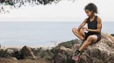 A woman in a vest and workout shorts sits on a rock outdoors, looking at a fitness tracker