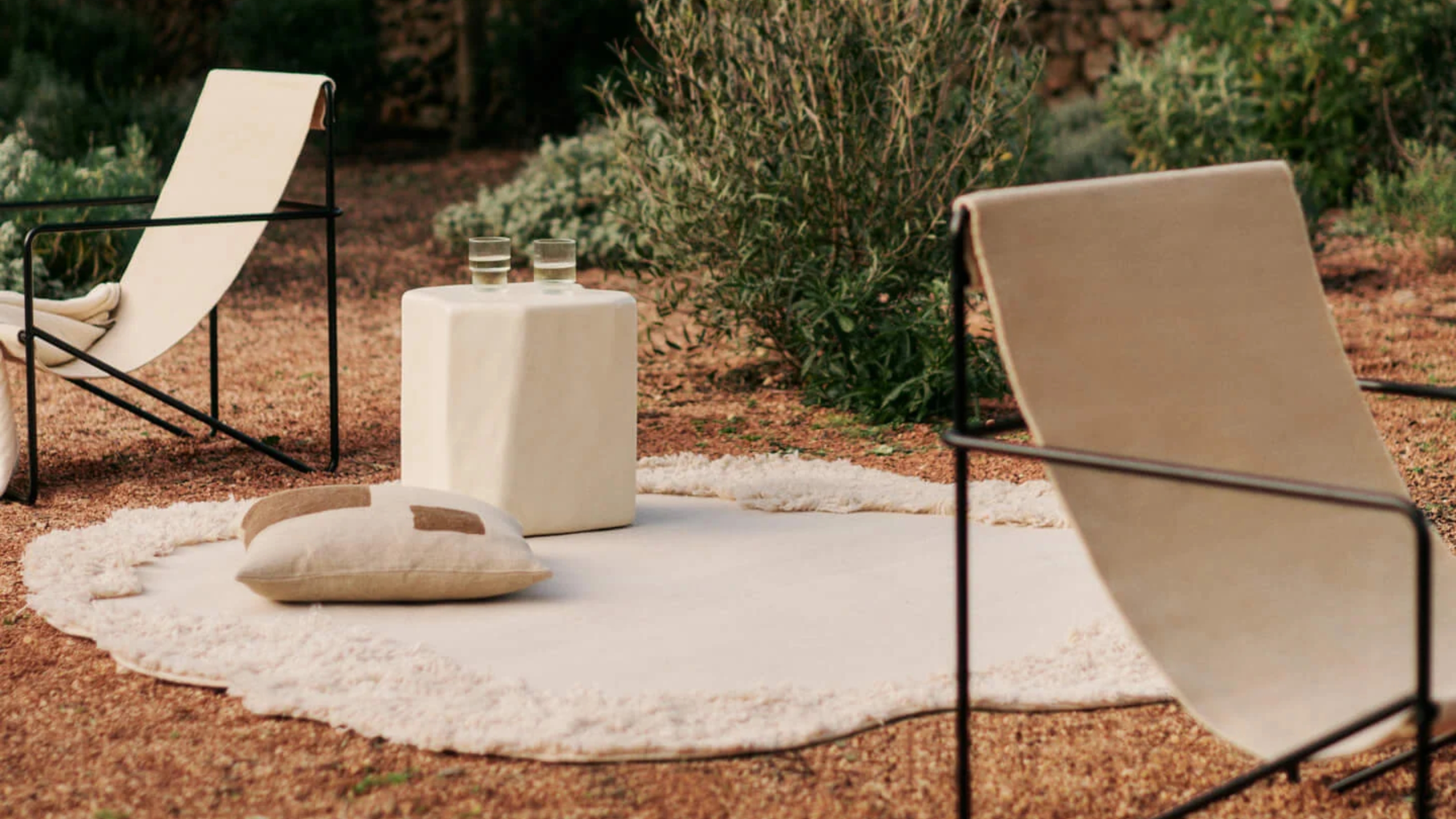 Image of a white, abstract-shaped outdoor rug with shaggy trim. It&#039;s lying on what looks like orange-toned mulch or pebbles. There is a white side table and a floor cushion on the rug, and two cream and black slingback chairs.