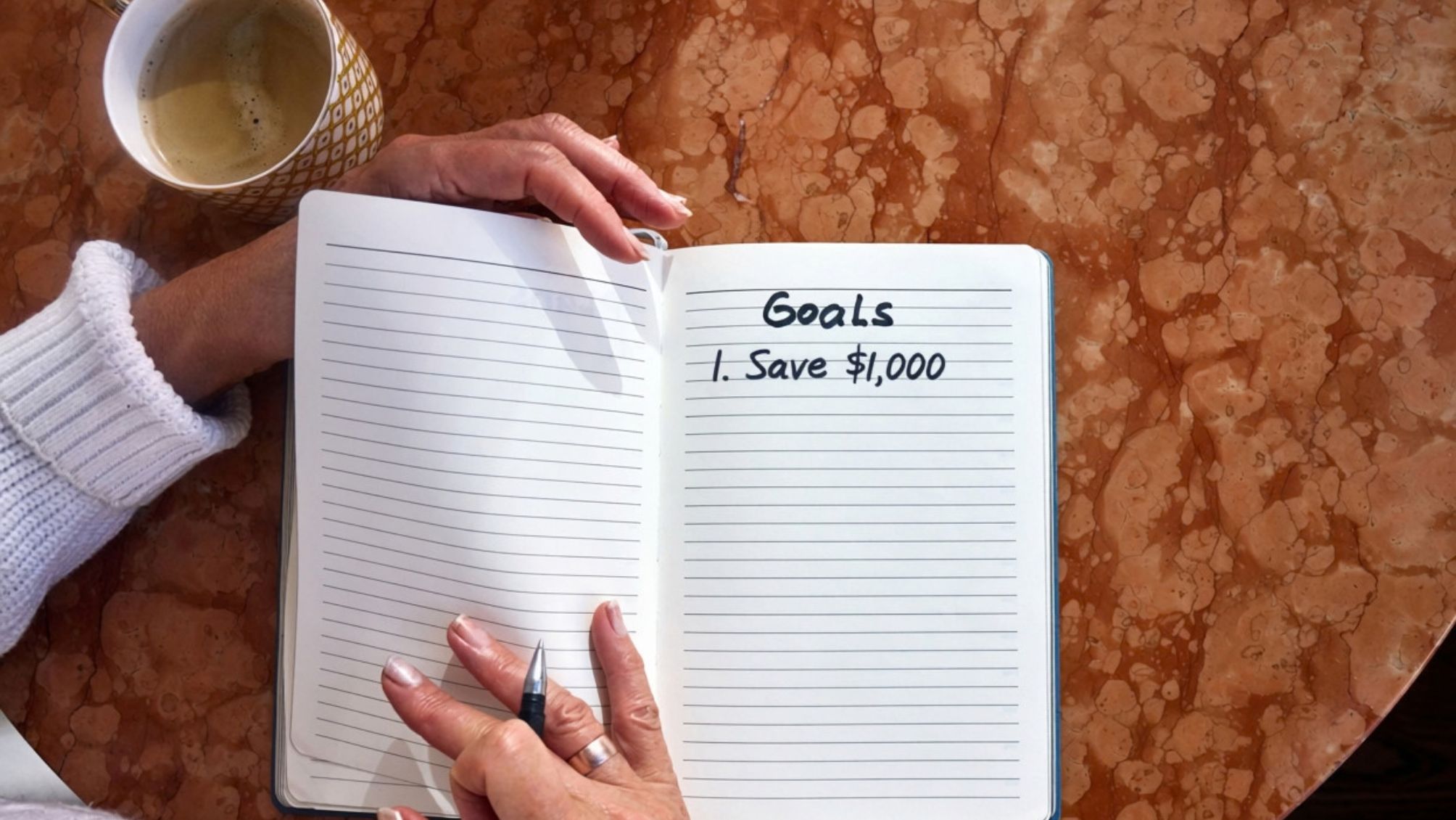 A person writing goals in a notebook. The first goal is to save $1,000