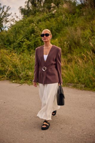 A woman wearing a brown cinched belted blazer, a white bralette, ivory maxi skirt, black sandals, black large sunglasses, and a black top-handle bag.