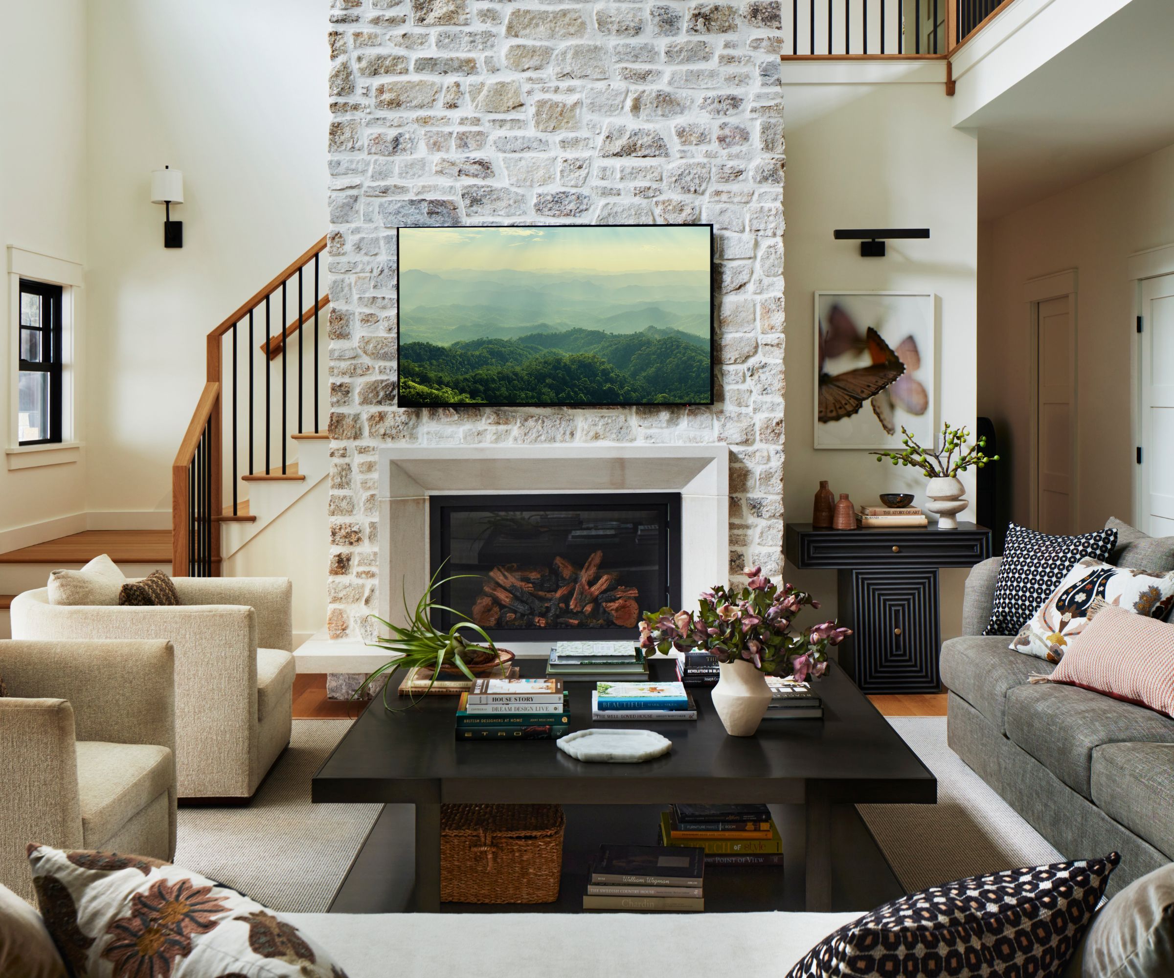 farmhouse style open plan living room with a large stone fireplace with frame TV, gray and beige sofas, soft yellow beige arm chairs and a black coffee table