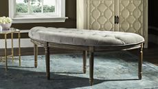 Button-tufted grey wooden bench pictured atop of a blue carpet inside of a sunlit living room alongside collected furniture