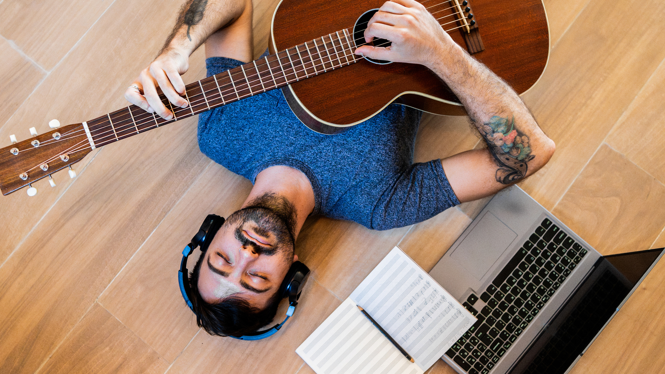 Ma in a blue t-shirt and headphone on lays on the floor playing an acoustic guitar