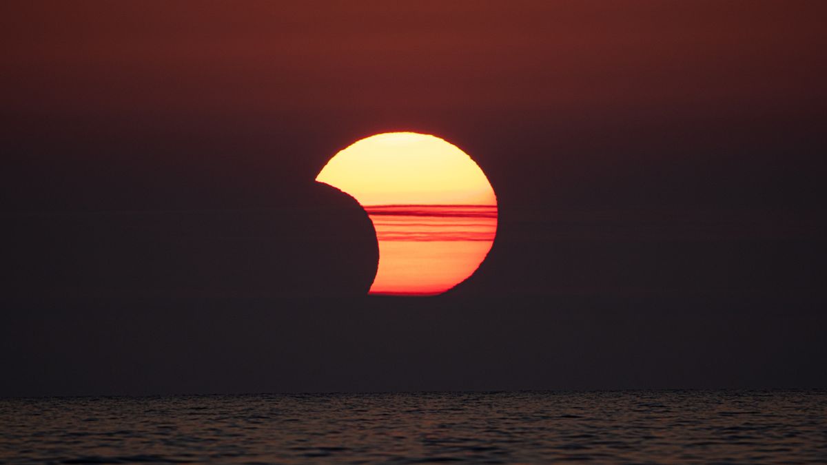 partial solar eclipse showing the moon appearing to take a &#039;bite&#039; out of the sun behind some hazy clouds above the ocean. 