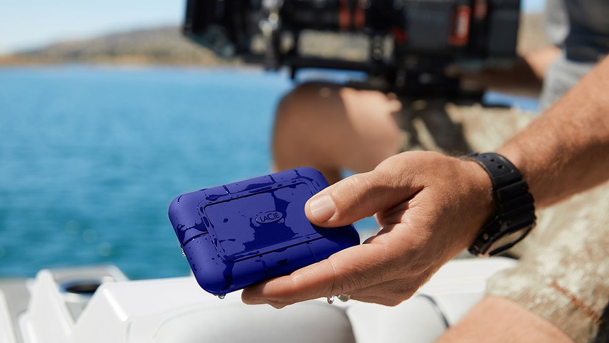 The LaCie Rugged SSD Pro5 held in a person&#039;s hands near a camera and water