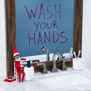an elf sat on the bathroom sink next to a bottle of hand soap, one of our favorite Elf on the Shelf ideas
