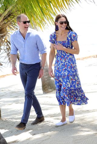 Kate Middleton wearing a blue floral dress and sunglasses walking across a beach with Prince William and laughing