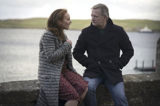 Meg (Lucianne McEvoy) and Perez (Douglas Henshall) sit on a low wall together, engaged in conversation. The sea is visible behind them.