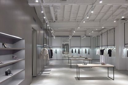 A fashion showroom featuring fashion accessories on the left, clothes items on hangers continuing around clothing on perimeter rails and fashion accessories on three tables in the centre. 