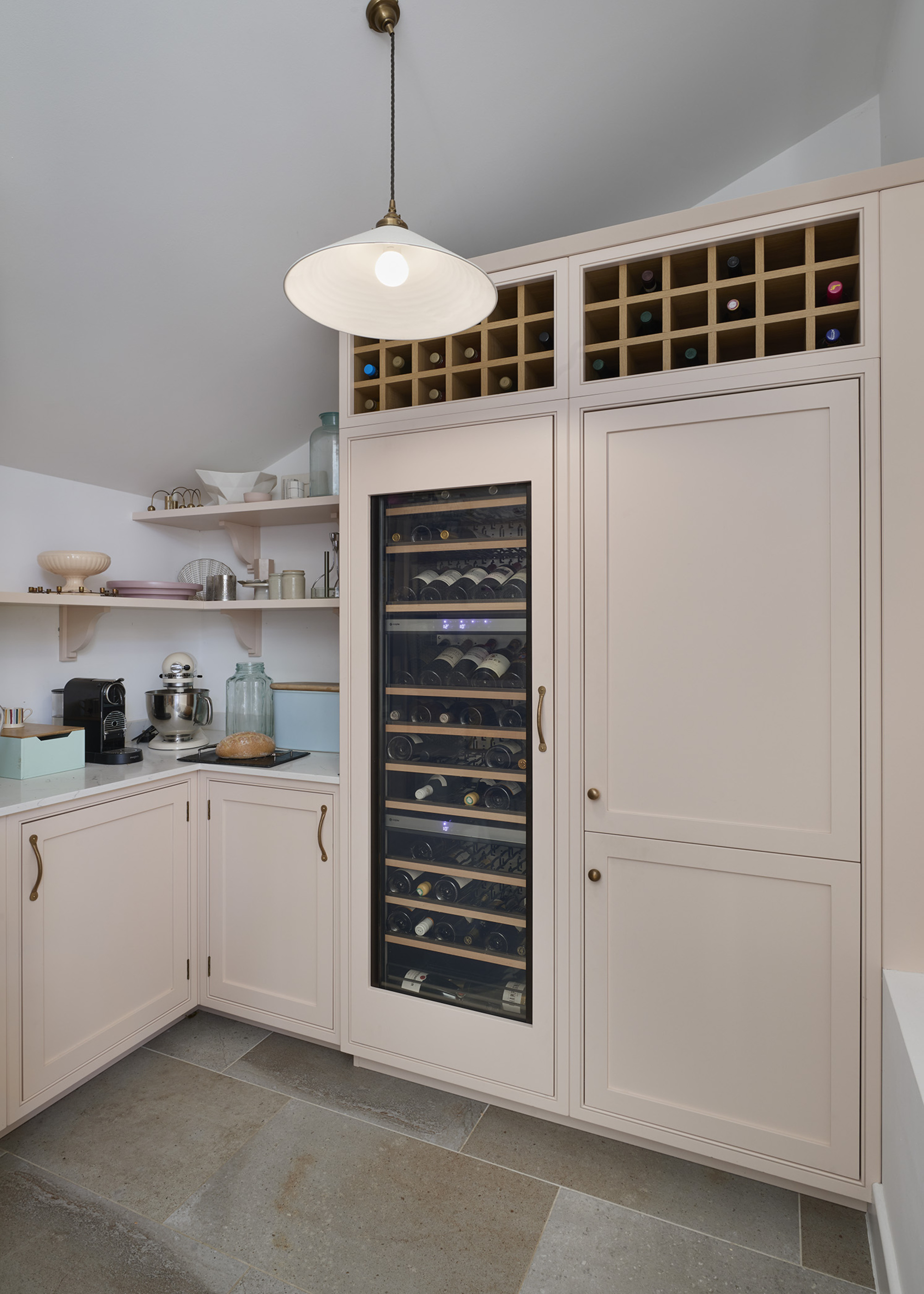 A kitchen with light pink cabinetry incorporating wine racks at the top, and a wine fridge with a glass window