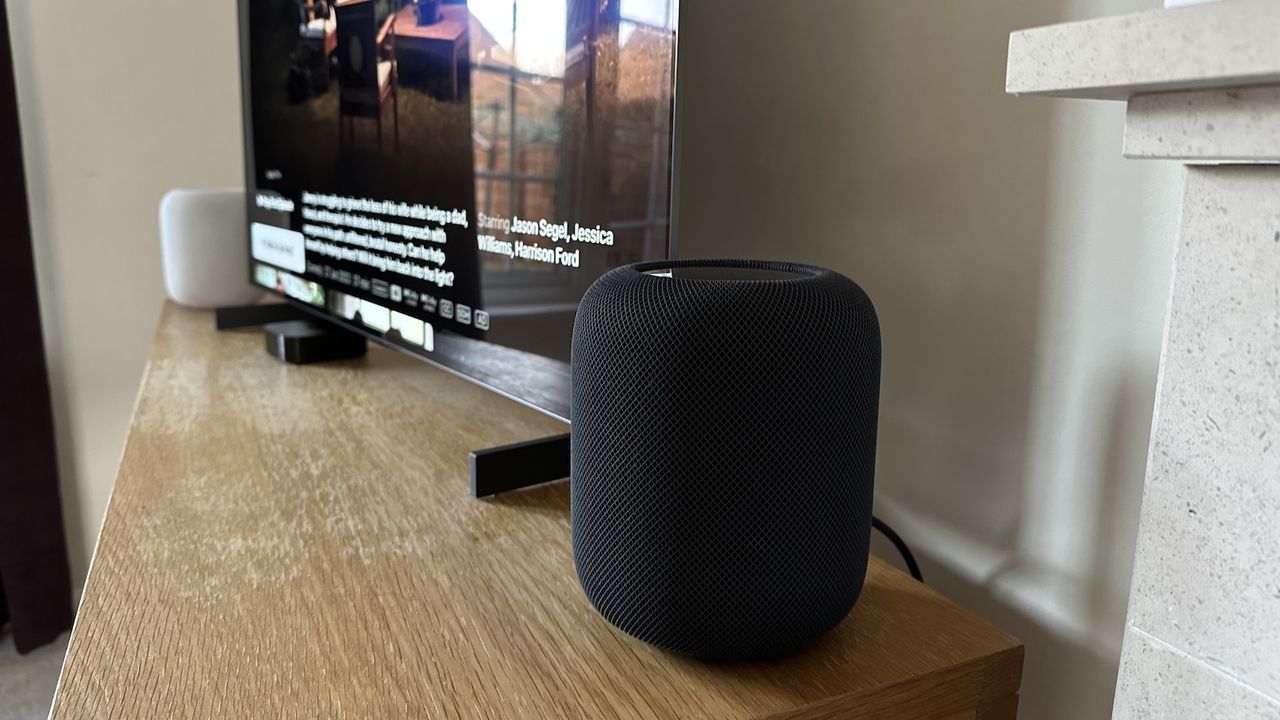 A dark grey Apple HomePod 2 smart speaker positioned next to a TV on a wooden surface. A second HomePod speaker in white is visible in the background, with the end of a mantelpiece just peeking into the foreground.