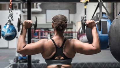 A woman in a sports bra holds dumbbell in a gym. She is facing away from the camera, so we see her back. Her arms are out to the side, with her elbows bent at right angles, so the dumbbells are in line with her head. In the background we see punching bags.