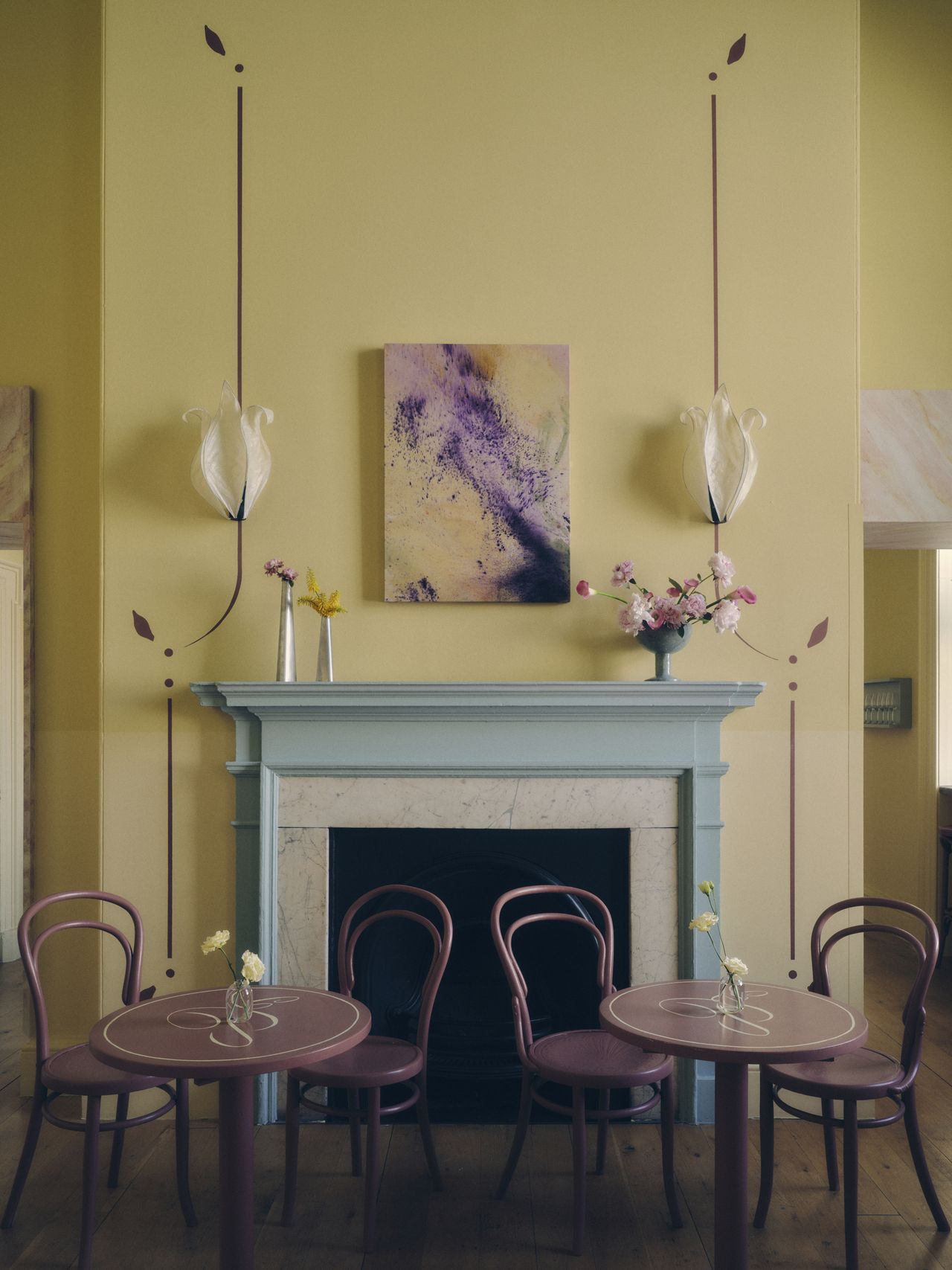 Image of a butter-yellow room with dark maroon/oxblood cafe tables and matching chairs. There is a light blue fireplace in the center wall with oxblood paint details on the yellow wall around it.