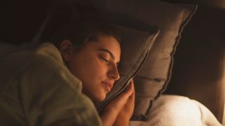 A close up photo of a young woman's face propped up on a pillow as she sleeps; her face is illuminated by a bedside lamp out of frame