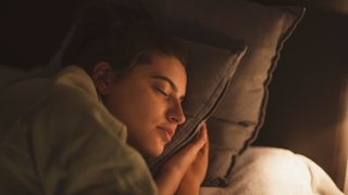 A close up photo of a young woman&#039;s face propped up on a pillow as she sleeps; her face is illuminated by a bedside lamp out of frame