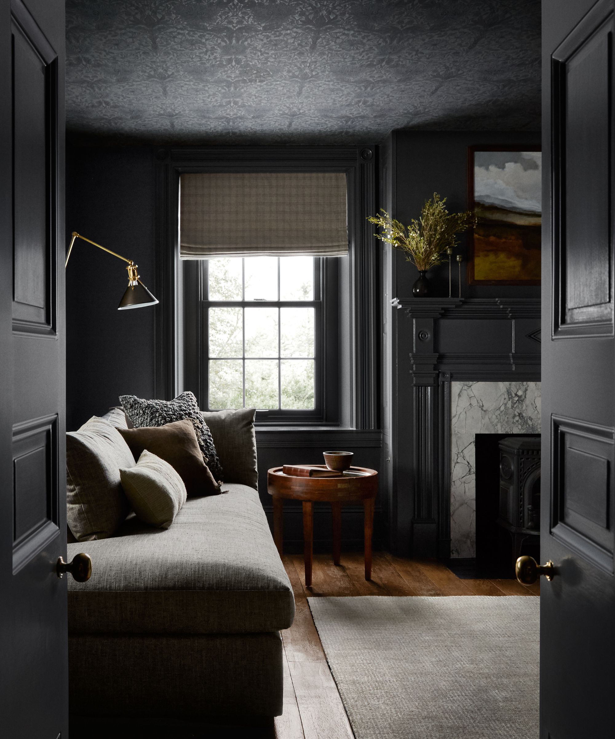 Dark living room with charcoal painted walls, wallpapered ceiling, a marble fireplace, wooden floors, a pale grey sofa, and a window with a plaid blind.