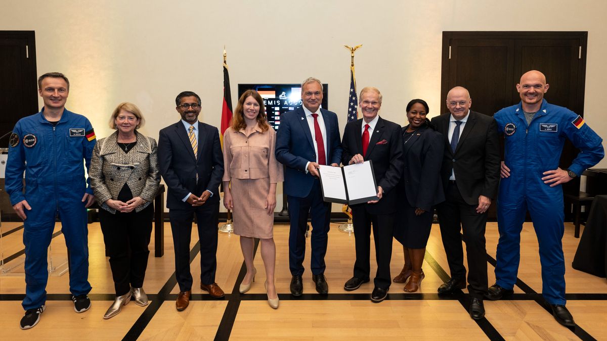 nine well-dressed people stand in a wood-floored room in front of the american and german flags.