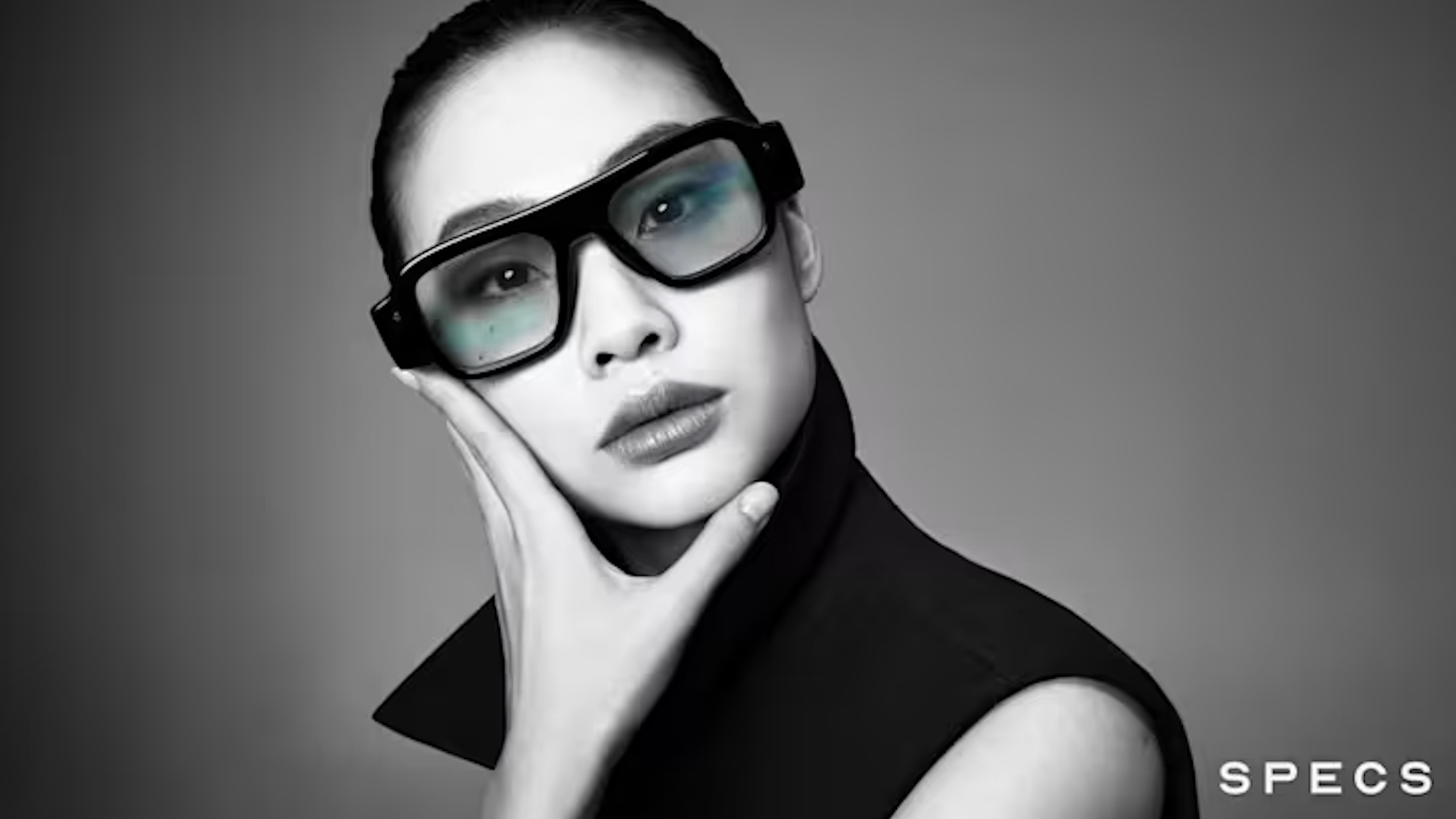 A black-and-white, close-up portrait of a woman with her hair pulled back, wearing thick, black-framed glasses with slightly tinted lenses and posing with her hand resting against her chin.