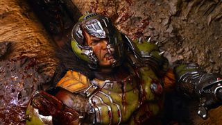 A warrior in a battle-scarred helmet and green armor lies on a gritty surface, covered in blood. His expression is defiant, with a futuristic and intense atmosphere.