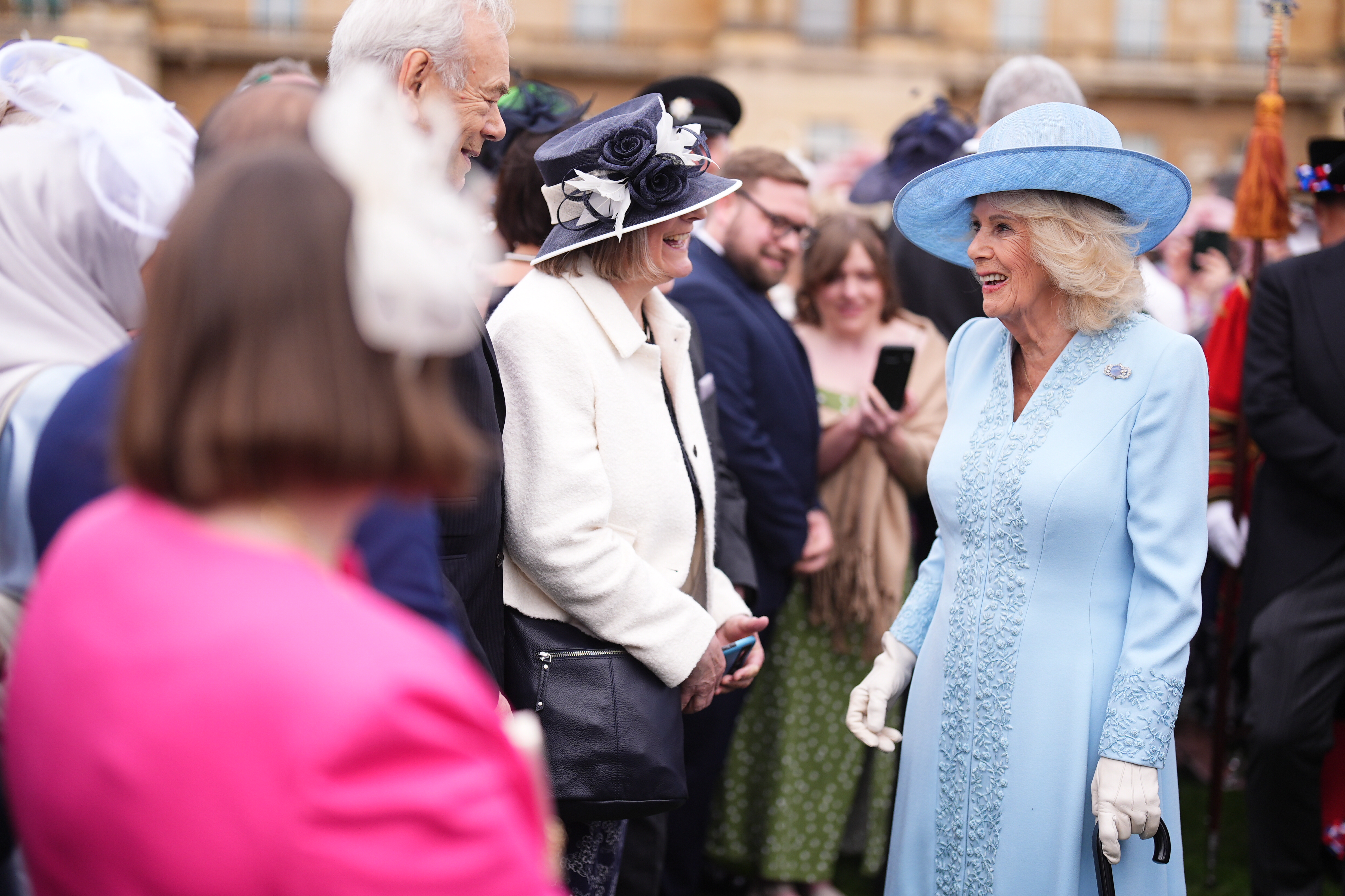 Queen Camilla wearing a blue coat and hat talking to people at a garden party