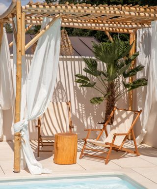 Two beige fabric and wooden sun chairs under a driftwood pergola with a curtaon on the side of it, by the swimming pool in warm sunlight