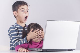 Two children using a computer with the older one cover the eyes of the younger one with an expression of shock