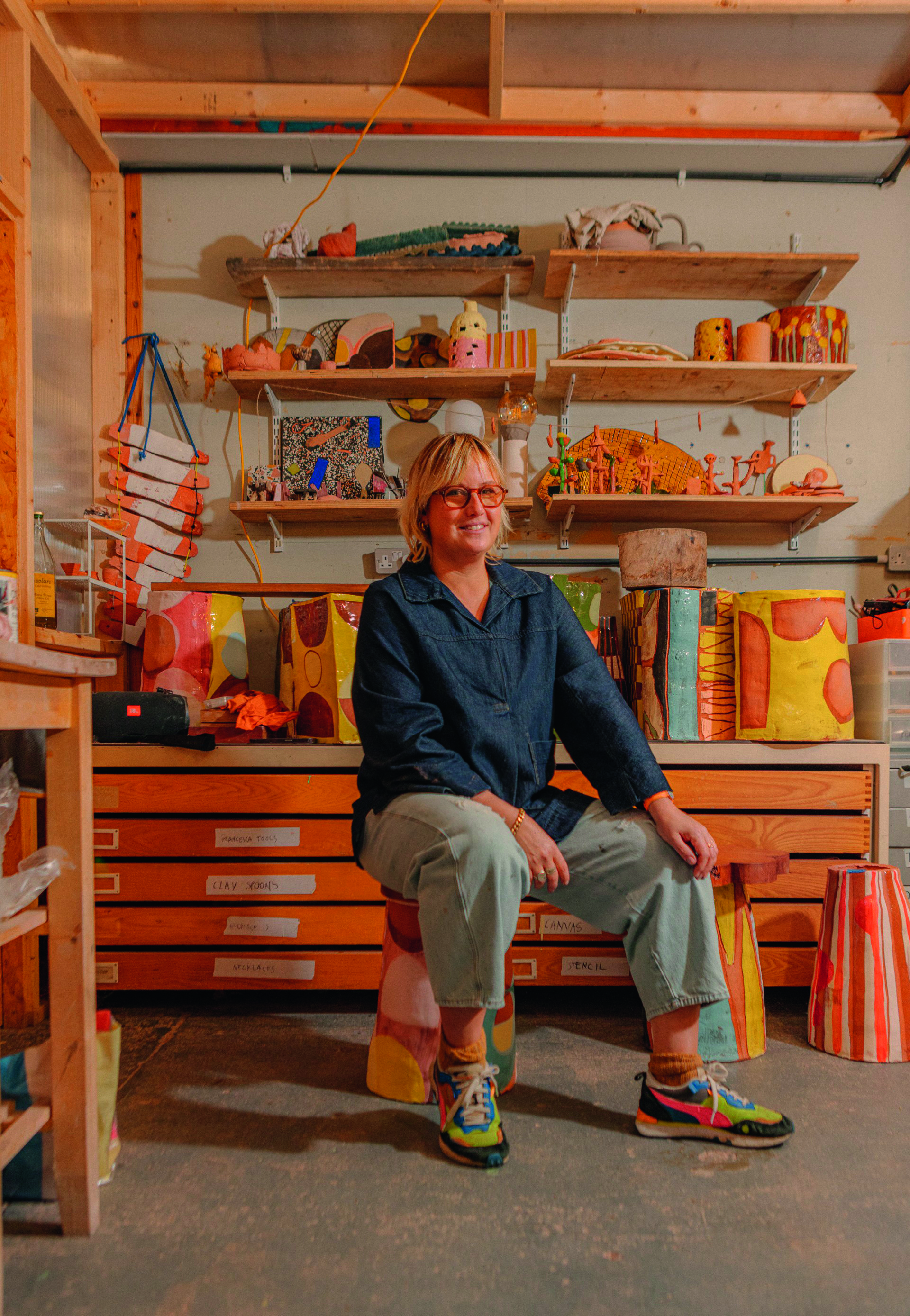 A young ceramist dressed in relaxed workwear in navy blue and grey, colorful sneakers, and red glasses sits on an eccentrically painted stool surrounded by equally striking creations in her studio.