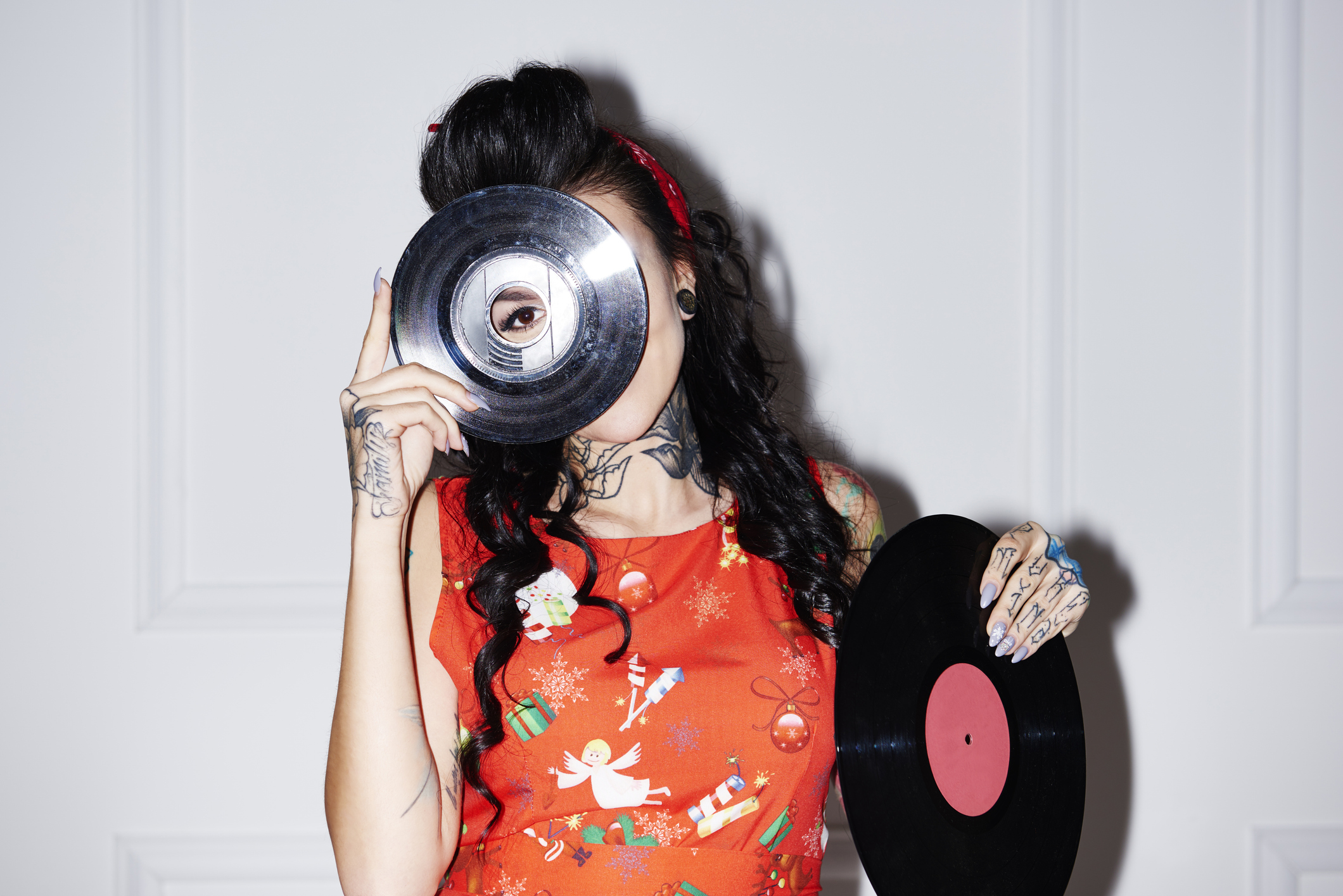 A woman dressed in vintage clothing holds a record over her face, with her eye peering through the record&#039;s center hole.