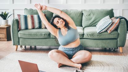 Curly haired young woman in top and shorts turns on online yoga training and practices exercises on floor mat against green sofa by wall