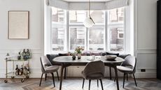 A bright dining space with a large modern table with soft brown chairs, and a gold bar cart to the left filled with bottles beneath a framed artwork. Behind the table is a cushioned bench seating area, and large bay windows.