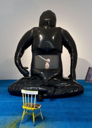 Black Inflated human like object sitting on a blue carpetted floor against a white wall with a tiny blue and yellow chain in front of it