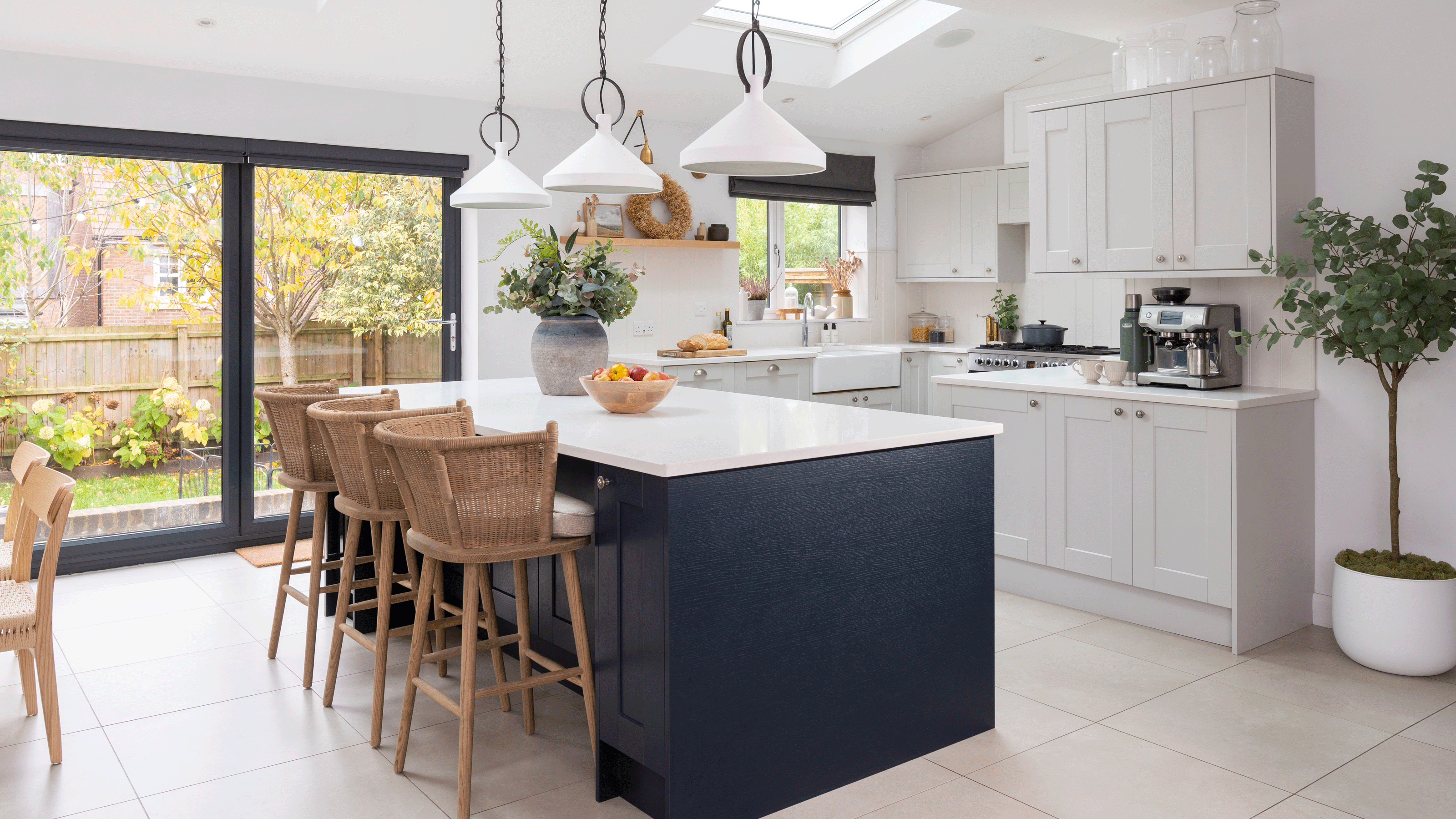 an open plan space with a white and black shaker kitchen and wicker bar stools and a Sage coffee machine beside a dining area with wooden dining furniture